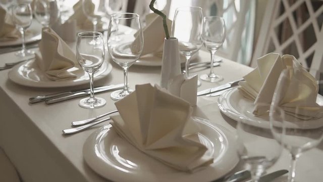Prepared table for a banquet, plates, cutlery, glasses and plants in vases. Waiting for guests in a celebration hall of restaurant in sunny day