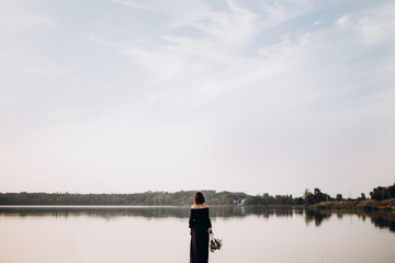 girl in a black long dress with a bouquet in hands stands near the water and looks at the sunset