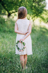 girl in full growth in a white dress from the back holds a wreath of pink roses and stands on a green garden