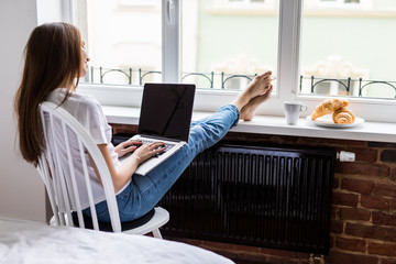 Relaxed young woman working laptop with legs on widnowsill with morning breakfast