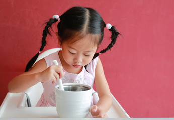 Close-up asian child girl eating pork bone soup on high chair against red wall background.
