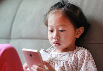 Little Asian girl playing smart phone and drinking milk with straw on sofa at home.