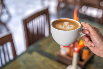Hot cappuccino latte coffee in white cup holding by hand in front of a table