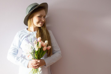 A young beautiful girl with blond dissolved long hair, a felt hat on her head, keeps spring flowers in her hands on a sunny day. Women's Day.