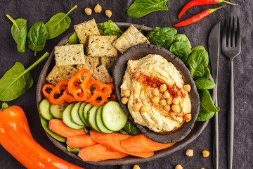 Homemade traditional hummus with vegetables, crackers on a black clay dish, dark background, top view. Healthy vegan food concept.
