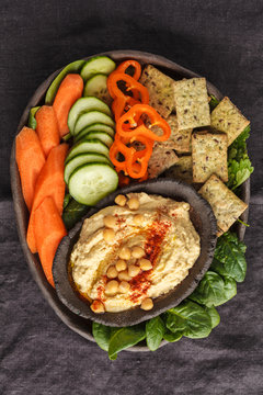Homemade traditional hummus with vegetables, crackers on a black clay dish, dark background, top view. Healthy vegan food concept.