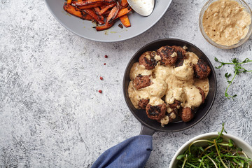 Flatlay with traditional swedish beef meatballs with gravy served in one portion black cast iron pan, handle wrapped in blue towel, plates with sauce, baked carrots and chickpeas. Dinner concept.