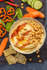 Homemade traditional hummus with vegetables, crackers on a black clay dish, dark background, top view. Healthy vegan food concept.