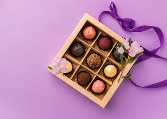 Assorted chocolates in a paper box with a satin purple ribbon on a bright background decorated with fresh flowers.
