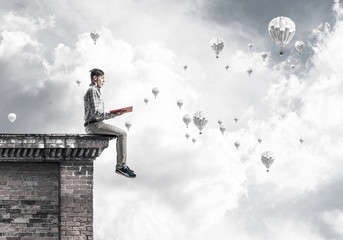 Man on roof edge reading book and aerostats flying in sky