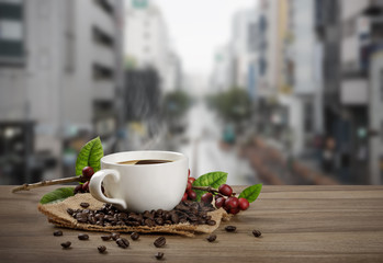 Hot coffee cup with fresh organic red coffee beans and coffee roasts on the wooden table and the city background
