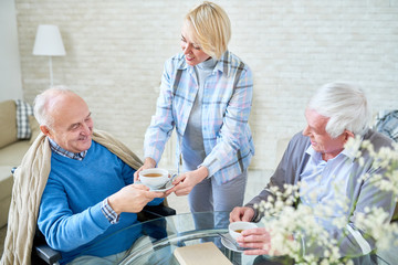 Portrait of blonde young woman serving tea to senior people  smiling happily while enjoying evening together in living room