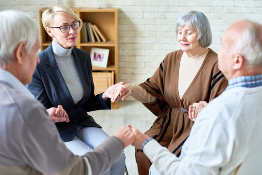 Senior people holding hands in group therapy session lead by female psychiatrist, focus on elegant elderly woman with eyes closed, copy space