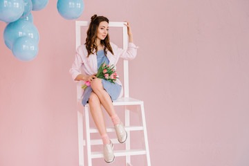 A girl in a blue dress and a pink shirt sits on a ladder in a pink room with a bouquet of tulips