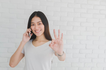 Asian beautiful girl using smartphone and showing okay in a white brick wall background.Smile face and feeling happy.
