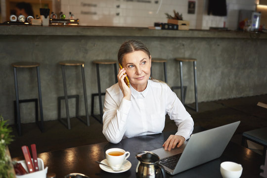 People, job, modern lifestyle, technlogy and communication concept. Picture of atractive confident mature businesswoman in formal wear having phone call durign coffee break at cafe and using laptop