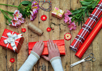 Festive background. Top view composition of woman hands wrap present for Birthday, Mother's day, Valentine's Day, March 8. Packed gifts and flowers, wrapping paper and tools on shabby wooden table