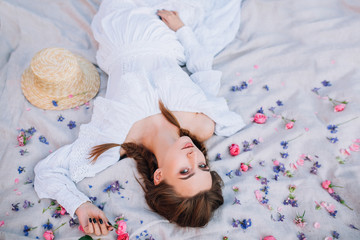 portrait of a pregnant girl in a white dress which lies on a gray fabric and holds a rose in her hand around which are scattered roses, peonies, wild flowers