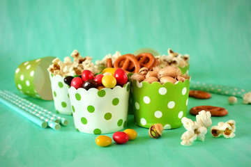 Popcorn, multicoloured drops, pretzels with salt and pistachio nuts in paper cups on green background. Snacks assortment