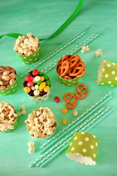 Popcorn, multicoloured drops, pretzels with salt and pistachio nuts in paper cups on green background. Snacks assortment