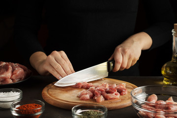 Female hands cut the raw meat into pieces on wooden board on dark table with seasoning and bottle of oil.