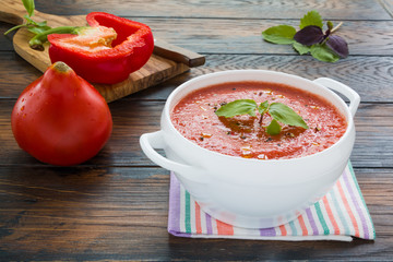 Cold soup gazpacho made with fresh tomatoes, garlic, bell pepper, onion and fresh basil leaves. White bowl on the rustic wooden table.