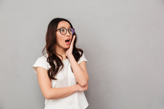 Shocked asian woman in t-shirt and eyeglasses holding her cheek