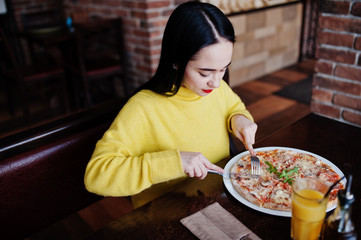 Funny brunette girl in yellow sweater eating pizza at restaurant.