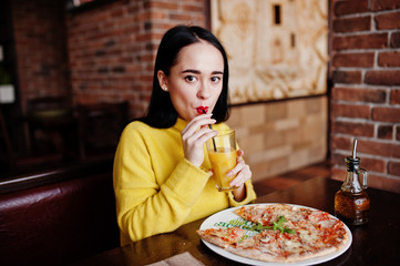 Funny brunette girl in yellow sweater eating pizza at restaurant.