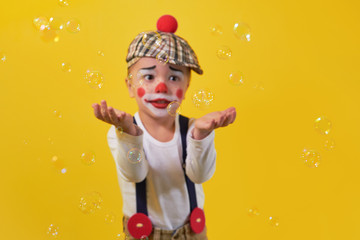Cute little boy clown in a costume, makeup, catches hands bubbles, soft focus. Kid on the bright yellow background. Clown looking to the soap bubbles. Concept birthday, holiday,celebration, party.