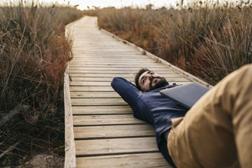 Adult man with laptop chilling on wooden pier in tall grass and enjoying freedom in sunlight.