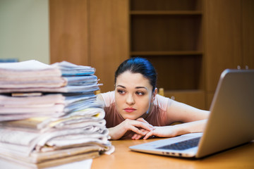 A young woman sits in front of a pile of papers and a computer holding her head