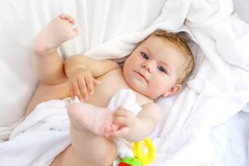 Obraz na płótnie Canvas Cute little baby playing with toy rattle and own feet after taking bath. Adorable beautiful girl wrapped in white towels