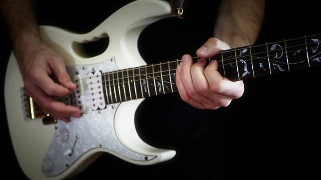 A man plays solo on a white electric guitar on a black background.