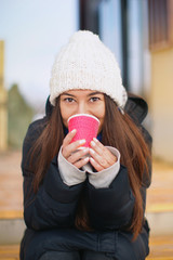 young beautiful woman drinks from a glass of coffee drink and tea in winter in a hat