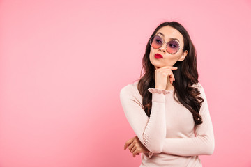 Photo of dreaming adult girl with red lips wearing trendy sunglasses looking aside touching chin with hand, isolated over pink background