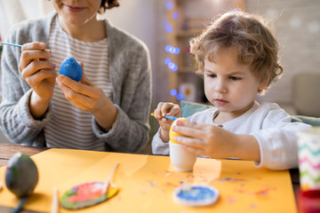 Portrait of adorable little boy painting Easter eggs making handmade decorations at home with mother, copy space