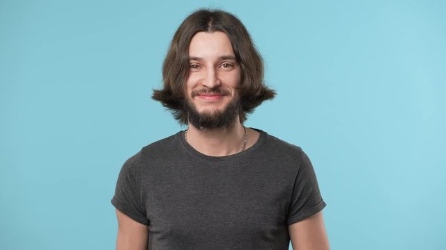 Portrait of young hairy man 20s wearing casual gray t-shirt and necklace smiling broadly in good mood, over blue background. Concept of emotions
