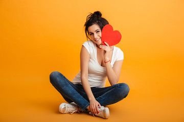 Photo of romantic teenage woman 20s in casual clothing sitting with legs crossed on the floor and holding paper red heart near face, over yellow background - Powered by Adobe