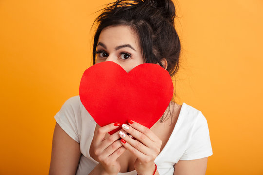 Closeup imege of gorgeous woman 20s in casual clothing covering her face with paper red heart and demonstrating affection, over yellow background