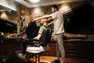 Obraz na płótnie Canvas Creating new hair look. Making haircut look perfect. Young bearded man getting haircut by hairdresser while sitting in chair at barbershop