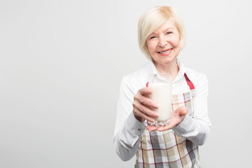 A picture where old woman wears blouse and apron and holding a cup of organic milk in her hands. She looks happy and delightful. Isolated on white background.