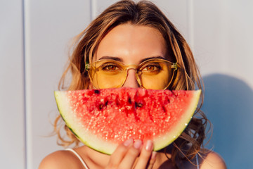 Charming lovely woman in yellow sunglasses hiding a half of her face with piece of watermelon,...