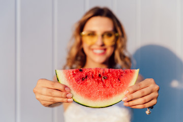 Blurred photo of charming joyful girl holding a piece of juicy watermelon with seeds, standing...