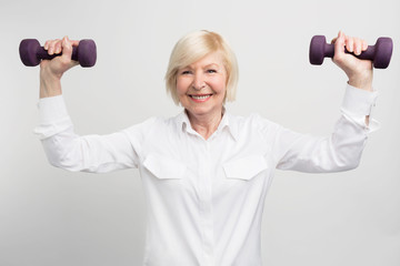 Cheerful madam is exercising with light weights. She does that because she has a careless retirement and much time to do that. Isolated on white background.