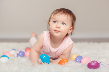 Cute adorable Caucasian baby girl in pink shirt lying on floor looking up and celebrating traditional Easter Christian holiday. Kid child holding and playing with colorful eggs