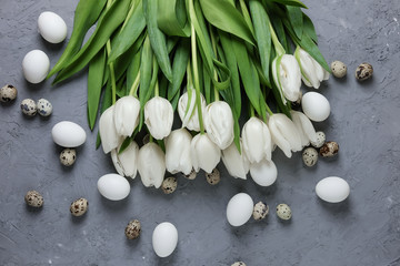 A bouquet of white tulips and chicken and quail eggs on a gray concrete background. Top view. Flat lay. Postcard for Easter and Spring Holidays.