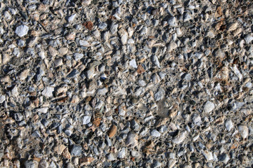 Gravel stones in the concrete. Different sized gravel stones are built into a concrete wall that is exposed to natural sunlight. Pebbles are mostly gray. In between are white, brown and black stones.