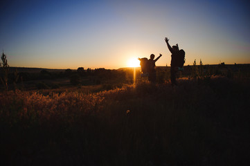 Fototapeta premium Silhouettes of two hikers with backpacks walking at sunset. Trekking and enjoying the sunset view