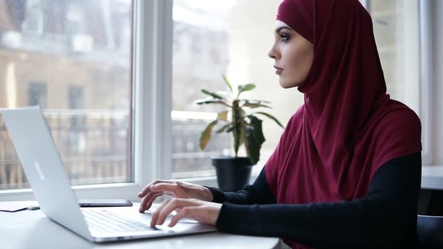 Young attractive supposedly muslim girl typing something on her laptop. Indoors footage